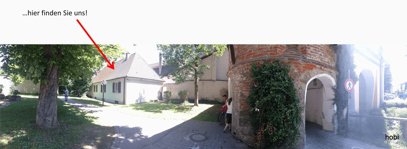Straßenfoto: Ein roter Pfeil mit dem Text „…hier finden Sie uns!“ zeigt auf ein helles, eingeschossiges Haus neben einem Torturm.
